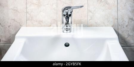 Robinet chromé avec levier pour mélanger de l'eau chaude et froide sur un lavabo blanc moderne dans une salle de bains avec des murs carrelés. Vie domestique et hygiène Banque D'Images