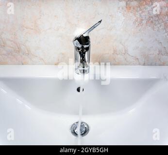 Robinet chromé avec levier pour mélanger de l'eau chaude et froide sur un lavabo blanc moderne dans une salle de bains avec des murs carrelés. Vie domestique et hygiène Banque D'Images