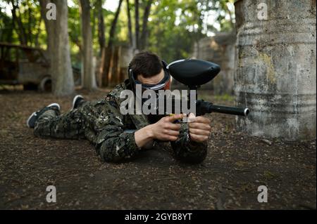 Un joueur de paintball masculin avec des pousses de fusil couché sur le sol, terrain de jeu dans la forêt en arrière-plan.Sport extrême avec arme pneumatique et peinture Banque D'Images