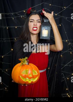 Joyeux Halloween ! Femme caucasienne tenant une grande citrouille sculptée dans un visage et une bougie lanterne stand devant le mur de décoration noir à halloween partie Banque D'Images