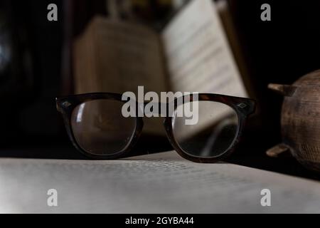 Vieilles lunettes usées reposant sur un livre ouvert avec des livres et une horloge en arrière-plan. Notion : temps de lecture Banque D'Images