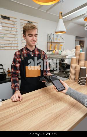 le barista souriant accepte le paiement depuis son téléphone portable via la machine de pos dans le café. le client tient son téléphone près du terminal nfc effectue le paiement mobile sans contact. Banque D'Images