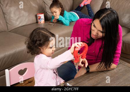 Mère encourageant la fille de 3 ans jouant avec des aimants et des objets métalliques, comme fille de 5 ans compte et trie des pièces de sa banque d'épargne en b Banque D'Images