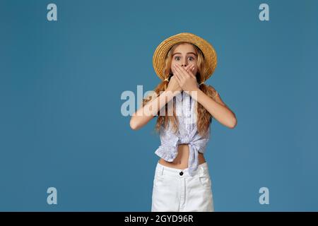 petite fille surprise en été chapeau couvre sa bouche avec ses mains sur fond bleu. Banque D'Images