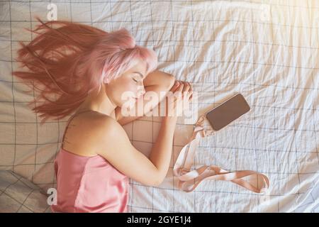 Magnifique modèle de femme endormi avec cheveux roses est avec téléphone mobile sur grand lit Banque D'Images