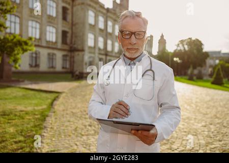 Portrait d'un médecin senior concentré en blouse de laboratoire et en lunettes tenant le presse-papiers, regardant à travers les notes, debout à l'extérieur près de la clinique Banque D'Images