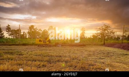 Paysage de champ de riz et forêt tropicale avec ciel de coucher de soleil. Pelle rétro travaillant en creusant le sol. Pelle hydraulique creusant sur le sol au champ de riz après la récolte Banque D'Images