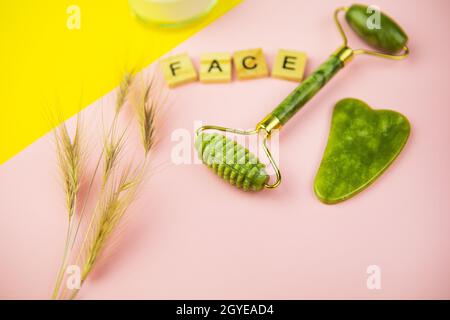 Outils de massage facial Gua Sha Green. Rouleau en jade de quartz vert sur fond rose-jaune. Pot de crème, inscription face en lettres de bois. CL Banque D'Images