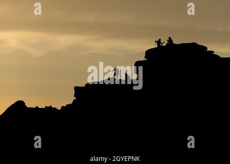 Personnes sur le bord de la Stanage, quartier de pointe, au coucher du soleil. Un couple prend un selfie tenant des boissons, silhoueté contre le ciel doré. Les grimpeurs sont là aussi Banque D'Images