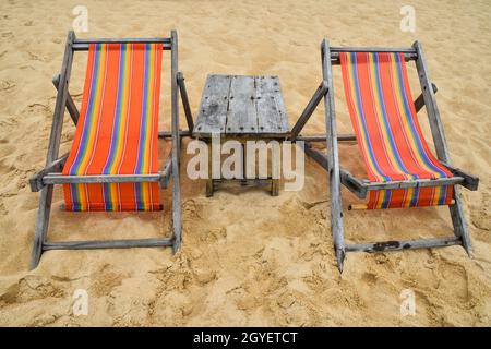 Deux chaises longues en toile colorées et vides et table en bois sur la plage de sable de la station balnéaire en Thaïlande, vue avant, grand angle Banque D'Images