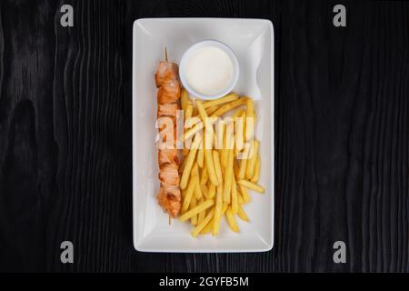 Shashlik au saumon grillé avec pommes de terre frites et sauce sur plaque blanche sur fond noir en bois Banque D'Images