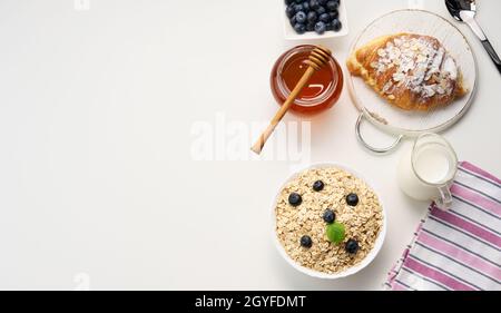 Petit déjeuner du matin, flocons d'avoine crus dans une assiette en céramique, lait dans un carafe, bleuets et miel dans un pot sur une table blanche, vue du dessus, espace copie Banque D'Images