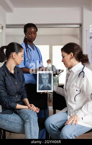 Radiologue spécialiste médecin analysant la radiographie clinique avec une patiente pendant un rendez-vous médical dans la salle d'attente de l'hôpital.Une infirmière afro-américaine écrit un traitement de santé sur presse-papiers Banque D'Images