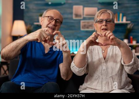 Couple âgé marié faisant le symbole du cœur avec les mains tout en regardant l'appareil photo dans le salon.Homme et femme à la retraite créant un signe d'amour romantique assis sur le canapé à la maison.Relation senior Banque D'Images