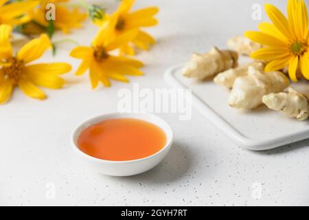 Sirop d'artichaut Jérusalem dans un bol, fleurs et racine sur fond blanc.Gros plan. Banque D'Images