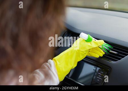Une fille en gants de caoutchouc jaune essuie le tableau de bord d'une voiture de la poussière avec une brosse spéciale lors d'un jour d'automne brillant.Mise au point sélective.Gros plan Banque D'Images
