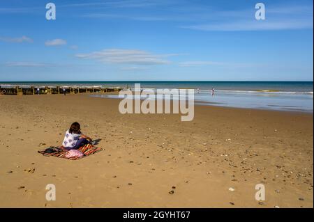 Femme d'âge moyen assise sur une couverture au soleil sur la plage de Mundesley presque vide, Norfolk, Angleterre. Banque D'Images