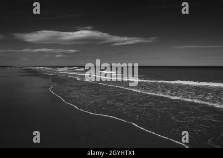 Photo noir et blanc sur le thème sombre d'une plage de sable avec vagues douces se brisant et nuages clairs sur l'horizon.Plage de Mundesley, Norfolk, Angleterre. Banque D'Images