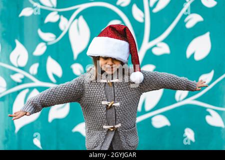 Petite fille portant le chapeau du père noël volant avec les bras ouverts comme un avion - drôle enfant jouant contre un fond de mur bleu à l'extérieur en période de Noël - Banque D'Images