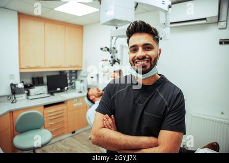 Une infirmière masculine de race mixte souriant avec les bras croisés debout dans la chambre du dentiste avec une collègue et un client de sexe féminin Banque D'Images