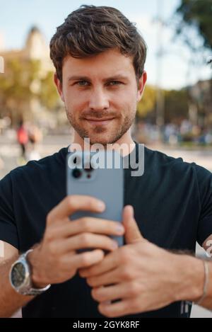 Auto-assuré jeune homme non rasé dans des vêtements décontractés prendre des photos sur un téléphone mobile moderne et regarder l'appareil photo tout en se tenant sur la place de la ville par beau temps Banque D'Images