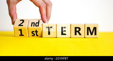 Symbole du premier ou du deuxième terme.Homme d'affaires tourne des cubes en bois et change les mots '1er terme' en '2e terme'.Belle table jaune, fond blanc.Busi Banque D'Images