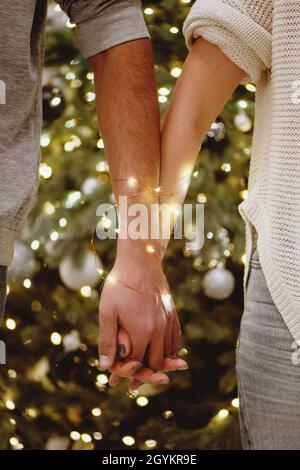 Vue partielle d'un couple tenant les mains enveloppées dans une guirlande avec arbre de Noël en arrière-plan. Banque D'Images