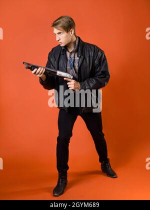 un combattant pour la justice, un gars dans une chemise décontractée et une veste en cuir avec un fusil de chasse, prêt à attaquer, photo studio sur fond orange Banque D'Images