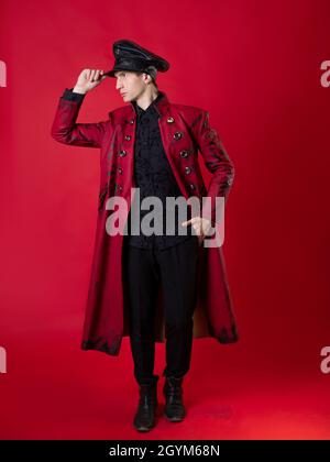 Un jeune homme scandaleux dans un manteau rouge audacieux et dans une casquette en cuir noir, style militaire, dans un style noir vintage, photo sur fond rouge Banque D'Images