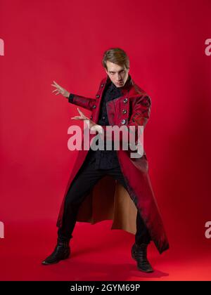 Un jeune homme scandaleux dans un manteau rouge audacieux dans un style noir vintage, pose épique et fait un geste de main. Photo sur fond rouge Banque D'Images