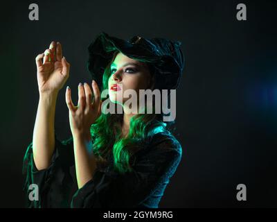 Une sorcière crée la magie, une jeune belle brune dans un chapeau pointu fait des passes avec ses mains, sorts. Portrait en gros plan, rétroéclairage vert Banque D'Images