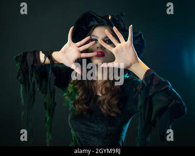 Une sorcière crée la magie, une jeune belle brune dans un chapeau pointu fait des passes avec ses mains, cache son visage avec ses mains. Gros plan portrait, galette Banque D'Images