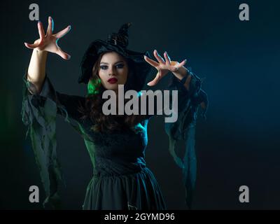 Une sorcière crée la magie, une jeune belle brune dans un chapeau pointu fait des passes avec ses mains, sorts. Fête du costume d'Halloween Banque D'Images