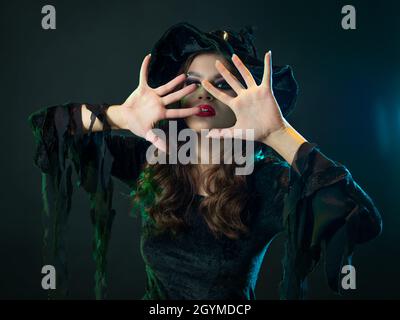Une sorcière crée la magie, une jeune belle brune dans un chapeau pointu fait des passes avec ses mains, cache son visage avec ses mains. Gros plan portrait, galette Banque D'Images