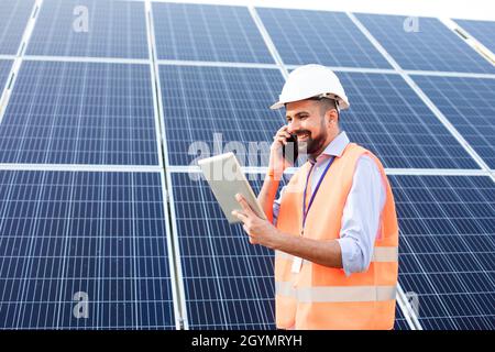 Le jeune électricien dans un gilet et un casque tient une tablette et parle au téléphone. Il se dresse sur fond de panneaux solaires Banque D'Images