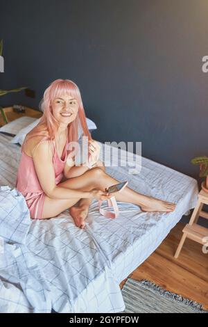 Portrait d'une belle femme aux cheveux roses et smartphone assis sur un grand lit Banque D'Images