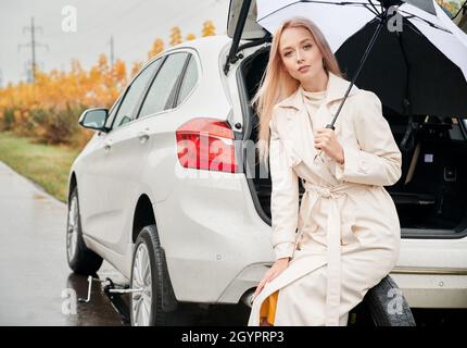 Portrait d'une jolie femme d'affaires blonde assise sur la roue de secours, tenant le parapluie de la pluie.Sur l'arrière-plan, voiture blanche avec coffre ouvert et buissons jaunes d'automne. Banque D'Images