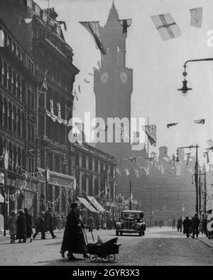 Une rue du centre-ville de Bradford, dans le Yorkshire, en Angleterre, bordée de drapeaux et de banderoles pour célébrer le couronnement du monarque britannique, le roi George VI, le 12 mai 1937.La tour de l'horloge toscane de 200 mètres ou le campanile de l'hôtel de ville de Bradford du XIXe siècle (anciennement l'hôtel de ville de Bradford) s'élève en arrière-plan.Au premier plan, une femme pousse un bébé à faible pente portant un petit drapeau d'Union ou drapeau d'Union, le drapeau du Royaume-Uni.Au-dessus d'elle, des drapeaux d'autres nations de l'Empire britannique volent aux côtés des Jacks de l'Union.L'hôtel de ville de Bradford a été construit en 1873 dans le cadre d'une conception primée par Lockwoo Banque D'Images