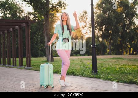 Vue sur toute la longueur du corps de la femme attrayante et gaie à cheveux gris, portant un sac aventure en faisant un salut à l'extérieur Banque D'Images