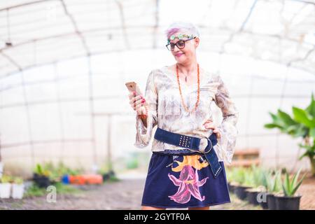 Femme âgée utilisant un téléphone portable tout en posant avec la main sur la hanche debout en serre.Une femme âgée rêvait d'être une messagerie texte hippie sur un téléphone portable.C Banque D'Images