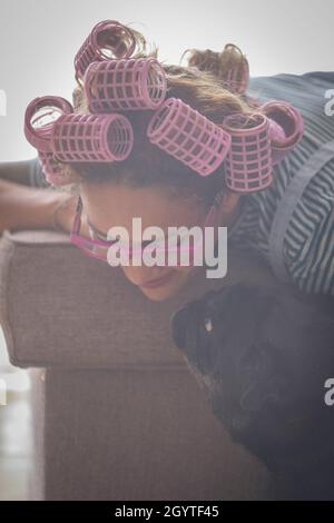 Jeune femme avec des curlers sur les cheveux et jouant avec son chien d'animal de compagnie se penchant sur le canapé dans le salon à la maison.Belle femme en lunettes de vue passant des loisirs t Banque D'Images