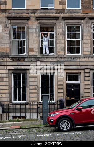 Commerçant sans équipement de sécurité debout sur un seuil de fenêtre au premier étage pour réparer les cadres de fenêtres sur une propriété à India Street, Édimbourg, Écosse. Banque D'Images