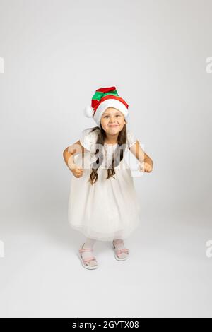 Portrait en longueur de fille gaie en robe blanche et chapeau de gnome de Noël dansant et appréciant des vacances, isolé sur fond blanc Banque D'Images