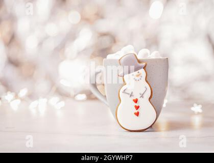 Biscuit au pain d'épice bonhomme de neige avec une tasse de chocolat chaud et des guimauves sur un fond chaud de lumières de Noël de bokeh. Banque D'Images