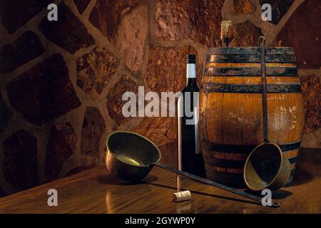 Ambiance vin.Un baril et deux louches de vin sur une table contre un mur de pierre.Au centre de la composition se trouve une bouteille noire avec une étiquette blanche. Banque D'Images