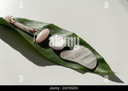 Grattoir de gouache rose, rouleau, pot de crème de feuille verte sur une table blanche. Équipement pour l'auto-massage et les soins de la peau pour le visage et le cou Banque D'Images