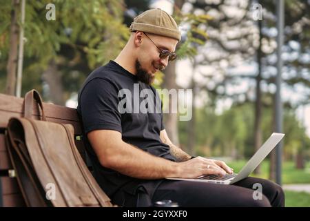 Photo extérieure du millénaire positif en lunettes de soleil qui se trouve sur un banc à l'ordinateur portable dans citypark Banque D'Images