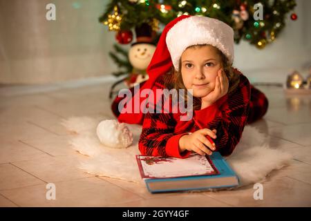 Une fille se trouve sur le sol, rêve et écrit une lettre au Père Noël, en arrière-plan il y a un arbre de Noël festif. Un enfant dans un chapeau de Santa Banque D'Images
