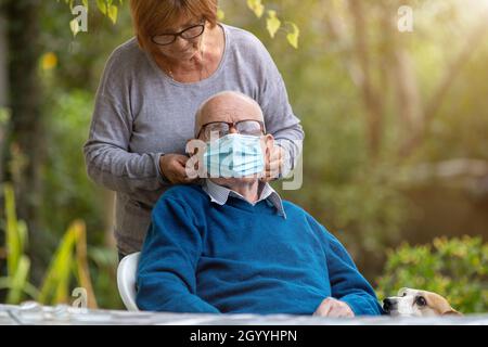 Femme mettant un masque de sécurité sur l'homme âgé dans le jardin tout en cuddling de chien mignon.Concept de protection contre les pandémies Banque D'Images