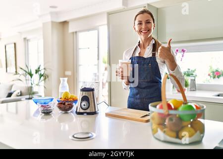 Belle femme blonde portant un tablier buvant smoothie à la cuisine souriant heureux et positif, pouce vers le haut faisant excellent et signe d'approbation Banque D'Images